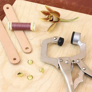 A Chicago screw tool with black rubber grips beside four brass Chicago screws, two punched leather straps and a spool of thread on a light wood surface.