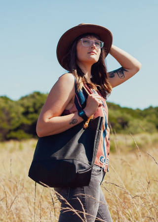 A woman in a wide-brim hat and glasses carrying a spruce and black Portsmith Tote with chestnut leather straps through a field of tall grass.